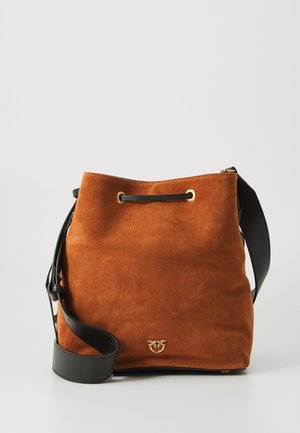 Bruine suède emmer-bag met zwarte leren schouderband en gouden metalen oogjes en logo-detail aan de voorkant tegen een effen achtergrond.