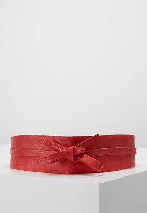 Wide red leather belt with a central knot detail, displayed on a white angled surface against a plain gray background.