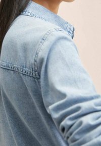 Chemise en denim bleu clair avec un col, des surpiqûres visibles le long des coutures et des manches retroussées, mettant en avant un tissu doux et texturé.