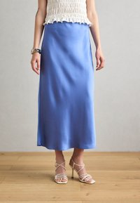 Satin tamnoplavi midi suknja s glatkom teksturom i ravnim krojem, u kombinaciji s bež sandalama s trakama.