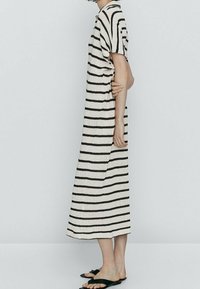 Persona che indossa un vestito midi a righe orizzontali bianche e nere e sandali neri, in piedi di lato con un braccio piegato dietro la schiena.