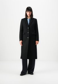 Donna con capelli scuri e lisci, che indossa un lungo cappotto nero, un top blu, pantaloni larghi scuri e scarpe nere, in piedi contro uno sfondo bianco semplice.