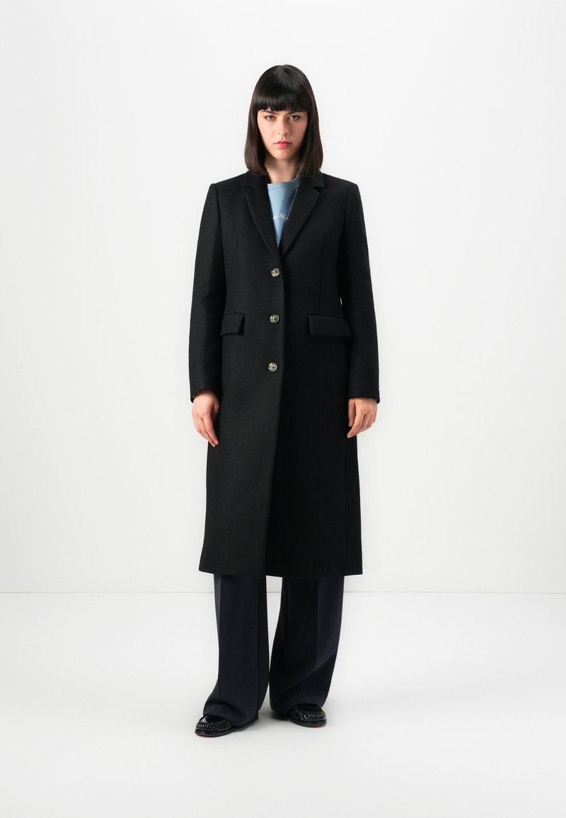 Donna con capelli scuri e lisci, che indossa un lungo cappotto nero, un top blu, pantaloni larghi scuri e scarpe nere, in piedi contro uno sfondo bianco semplice.