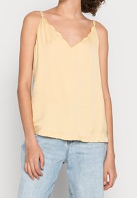 Gul, ärmlös topp med en vågig halsringning och tunna axelband; tillverkad av ett lätt tyg med en något skrynklig textur.