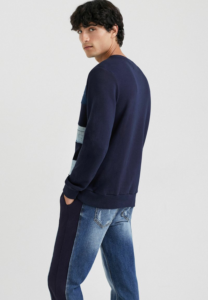 Sweatshirt à col rond de couleur marine avec des accents rayés bleu clair, en tissu doux. Assorti à un jean en denim bleu délavé avec un léger aspect usé.