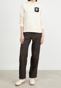 Sudadera con capucha color crema con un logo de letra "G" en negro, combinada con pantalones negros con detalles de costuras y zapatillas blancas con suelas beige.