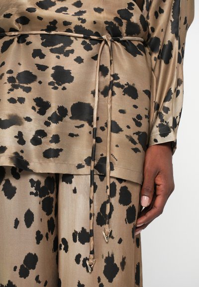 Beige top with black splatter patterns, featuring a tie waist with metal accents. Smooth, shiny fabric; long sleeves, relaxed fit.