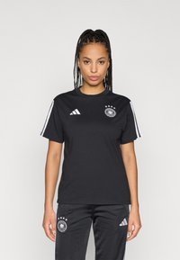 Fonott hajú nő fekete Adidas sportruházatban, német labdarúgó csapat logójával a pólón és a nadrágon, egy sima háttér előtt áll.