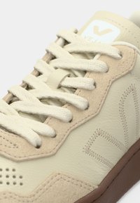Primer plano de zapatilla Veja beige con cordones tejidos, paneles de ante y cuero, logotipo "V" cosido y suela de goma marrón.