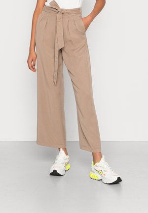 Calças largas beje com uma cintura cingida, apresentando pregas e bolsos laterais. Combinadas com sapatilhas brancas com detalhes em néon.