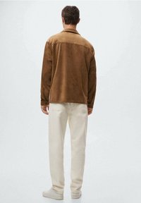 Brun ruskinds skjorte med krave, lange ærmer og lige snit, parret med off-white bukser og lyse sko.