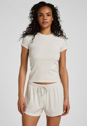 Vrouw met krullend haar, gekleed in een aansluitend lichtbeige topje met korte mouwen en bijpassende short met trekkoord, staand tegen een effen achtergrond.