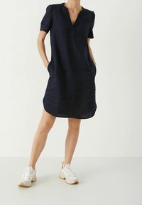 Nainen, jolla on päällään lyhythihainen tummansininen mekko ja valkoiset urheilukengät, seisoo kädet taskuissa vaaleaa taustaa vasten.