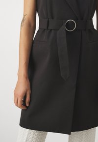 Černé bezramínkové šaty s páskem v pase se stříbrnou kruhovou sponou, bočními kapsami a strukturovaným designem, doplněné o texturované bílé kalhoty.