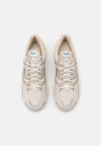 Beige athletic shoes with mesh and synthetic upper, featuring a cushioned sole, lace-up front, and subtle green logo accents.