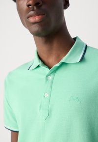 Polo verde con tejido texturizado, tres botones y detalles en azul marino en el cuello. Sutil logo de la marca bordado en el pecho.