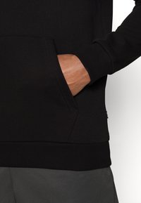 Czarna bluza z kieszenią z przodu, ściągaczami przy rękawach i u dołu. Tkanina sprawia wrażenie gładkiej, z matowym wykończeniem. Posiada metkę z logo z boku.