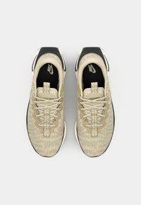 Beige Nike sneakers har en struktureret mesh-overdel med et bølget mønster, hvide snørebånd, en sort sål og logo-detaljer på tungen og siden.