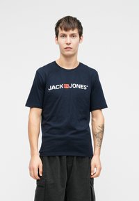 Námorně modré bavlněné tričko s krátkým rukávem. Na přední straně je bílo-oranžové logo JACK & JONES. Model má na sobě tmavé kalhoty.