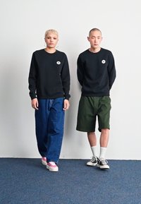 Sudaderas negras con parches de logo, jeans de mezclilla azul, pantalones cortos verdes y varios estilos de zapatillas; diseño simple con colores neutros.