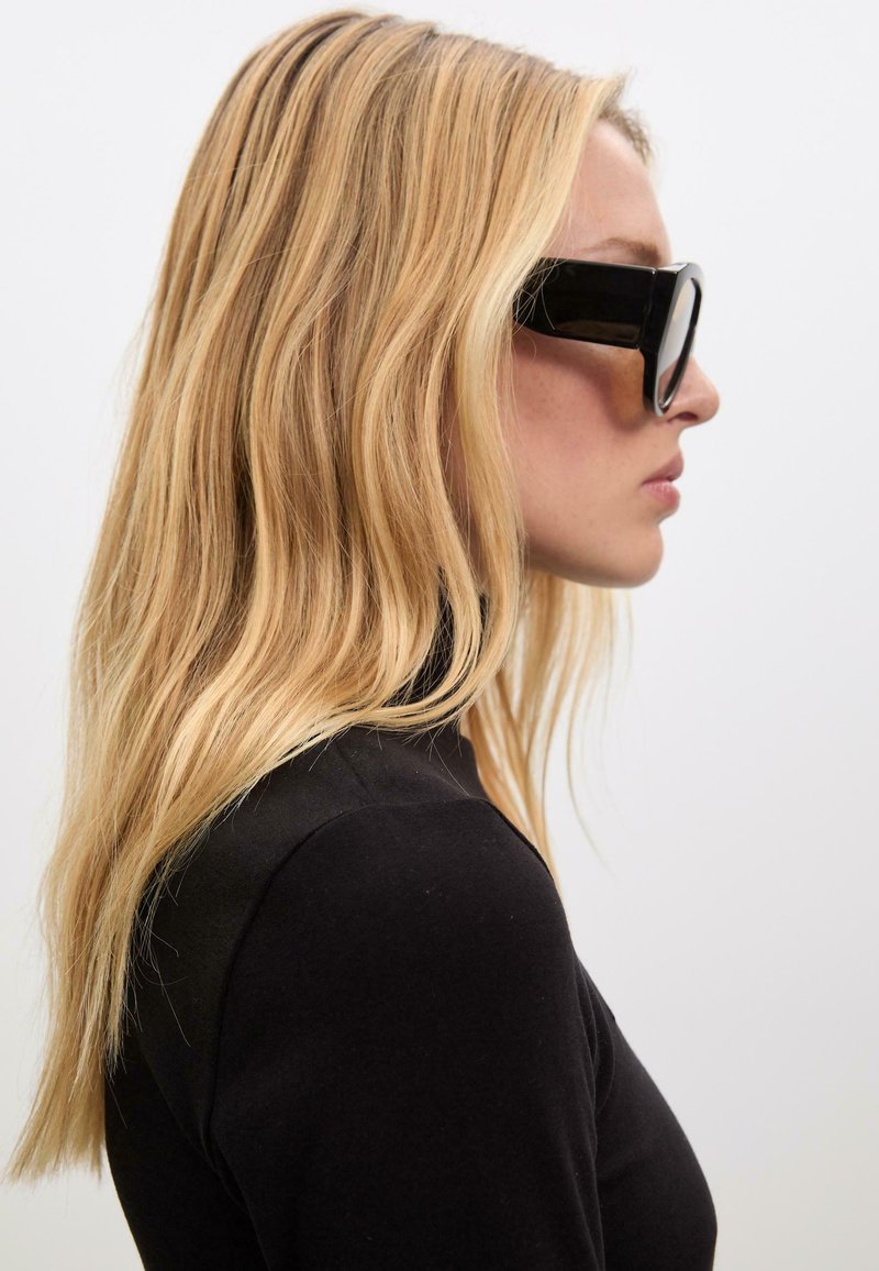 Femme blonde en col roulé noir portant de grandes lunettes de soleil noires, vue en profil sur un fond clair et uni.