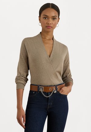 Woman wearing beige long-sleeve V-neck top, dark blue jeans, brown belt with silver chain and circular charm, standing with one hand in pocket.