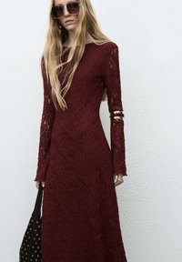 Burgundy lace dress with long sleeves, textured pattern, and gold accent bracelets. Black bag with grommet detailing. White background.