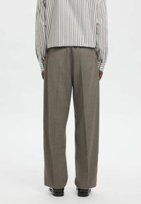 Oseba, oblečena v sive hlače širokih nogavic, belo in sivo dolgo srajco z navpičnimi črtami ter črne čevlje, stoji obrnjena stran na belem ozadju.