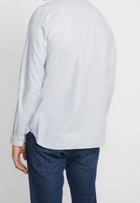 Světle modrá košile s dlouhým rukávem a texturovaným povrchem, zakulaceným spodním lemem a knoflíkovými manžetami. Nositá přes tmavě modré džíny.