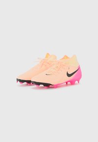 Des crampons de football Nike avec une tige de couleur pêche clair, des accents noirs et une semelle rose dotée de six crampons, conçus pour une traction sur les surfaces en gazon.