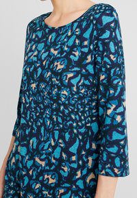 Rundhalsad topp med trekvartslånga ärmar i mörkblått med turkos, svart och beige abstrakt mönster. Mjuk, texturerad tyg.