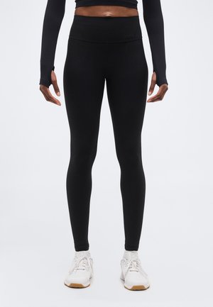 Persoon draagt zwarte leggings met hoge taille, een zwart topje met lange mouwen en duimgaten, en witte sportschoenen, staand tegen een effen achtergrond.