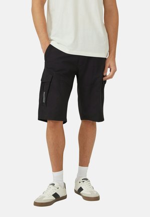 Mężczyzna w czarnych krótkich spodenkach cargo, białych skarpetach do kostek, białych i czarnych sneakersach oraz białej koszulce, stojący z rękami w kieszeniach.