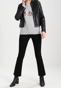 Svart läderjacka över en grå tröja med röd fågelbroderi, matchad med svarta utsvängda byxor och svarta ankelstövlar.