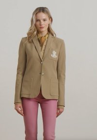Blazer beige avec deux poches avant et un logo brodé blanc. Associé à un pantalon rose et une chemise jaune à motifs. Tissu lisse.