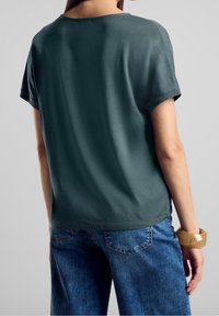Haut vert foncé à manches courtes avec un col rond, texture lisse et coupe décontractée, associé à un jean en denim bleu et un bracelet beige.