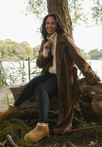 Uśmiechnięta kobieta w brązowym płaszczu, polarowej kurtce, czarnych legginsach i beżowych butach, siedzi na korzeniach drzewa nad jeziorem, z zielenią w tle.