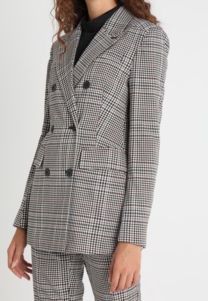 Vrouw draagt een zwart-wit geruite, dubbelgeknoopte blazer met bijpassende broek en een zwart topje eronder.