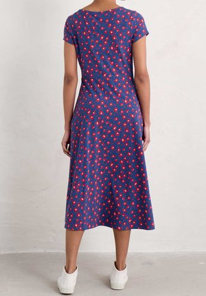 Femme portant une robe midi bleue à imprimé floral rouge et manches courtes, debout pieds nus sur un sol gris clair contre un mur blanc.