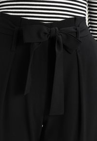 Svarta byxor med en mjuk textur, med ett knutet tygbälte i midjan och en hög midja. Randig topp delvis synlig.