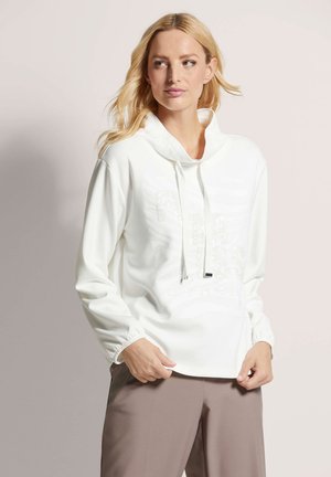 Sweatshirt blanc à col montant, avec cordon de serrage et poignets élastiques, présentant un subtil motif texturé sur le devant. Associé à un pantalon beige.