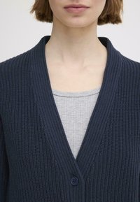 Gilet tricoté bleu marine avec un col en V, doté d'une fermeture à un bouton, d'une texture côtelée, porté par-dessus un haut côtelé gris clair.