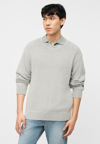 Νέος άνδρας που φοράει ανοιχτό γκρι πλεκτό πουλόβερ πάνω από πουκάμισο με γιακά σε απόχρωση φασκόμηλου και ανοιχτό μπλε τζιν, κοιτάζει προς τα αριστερά του μπροστά σε λευκό φόντο.