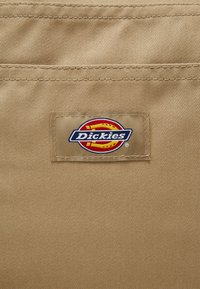 Beige kangas, jossa on vaakasuorat tekstuurit, esittelee suorakulmaista logo-patchia, jossa lukee "Dickies" valkoisella punaisella ja keltaisella soikealla taustalla.