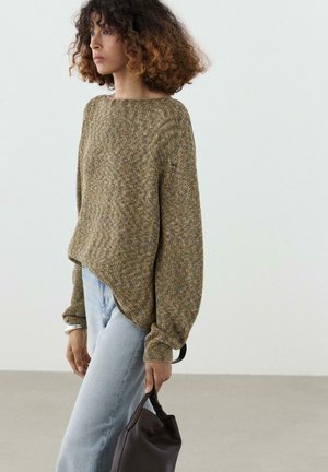 Kobieta z kręconymi włosami w luźnym oliwkowozielonym swetrze z dzianiny i jasnoniebieskich dżinsach, trzymająca czarną torebkę przy boku.