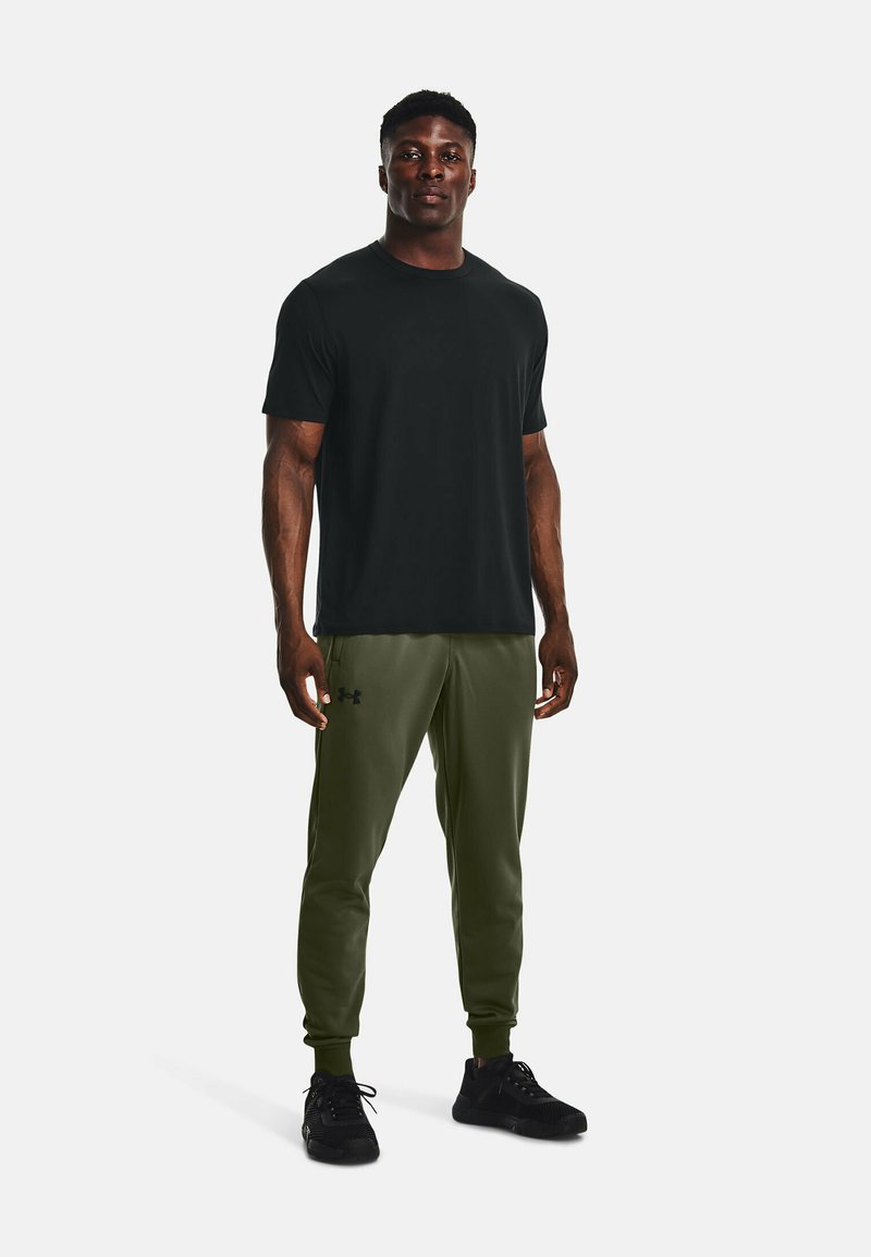 T-shirt noir à manches courtes associé à un jogger vert olive avec une taille élastique et un logo noir en accent. Des chaussures de sport noires complètent la tenue.
