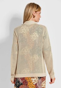 Beige gestrickter Cardigan mit loser, offener Maschenstruktur; mit einem Kragen und langen Ärmeln, kombiniert mit einem gemusterten Rock.