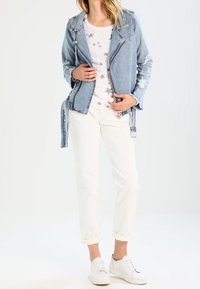 Kvinna iklädd en sliten denimjacka, en vit topp med stjärnmönster, vita uppvikta byxor och vita sneakers som står med händerna justerande jackan.