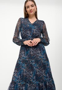 Langarm jurk van marineblauwe doorschijnende stof met een bloemenpatroon in blauw, wit en bruin. Bevat een gelaagde rok en rimpelige manchetten.
