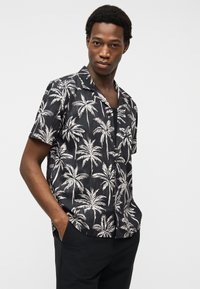 Νέος άνδρας που φορά μαύρο κοντομάνικο πουκάμισο με λευκές εκτυπώσεις φοίνικα, με τα χέρια στις τσέπες, στέκεται μπροστά σε ένα απλό φωτεινό φόντο.